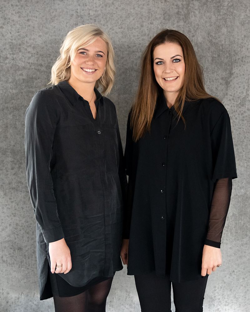 Christina og Camilla fra Salon Jyderne smiler foran en betonvæg